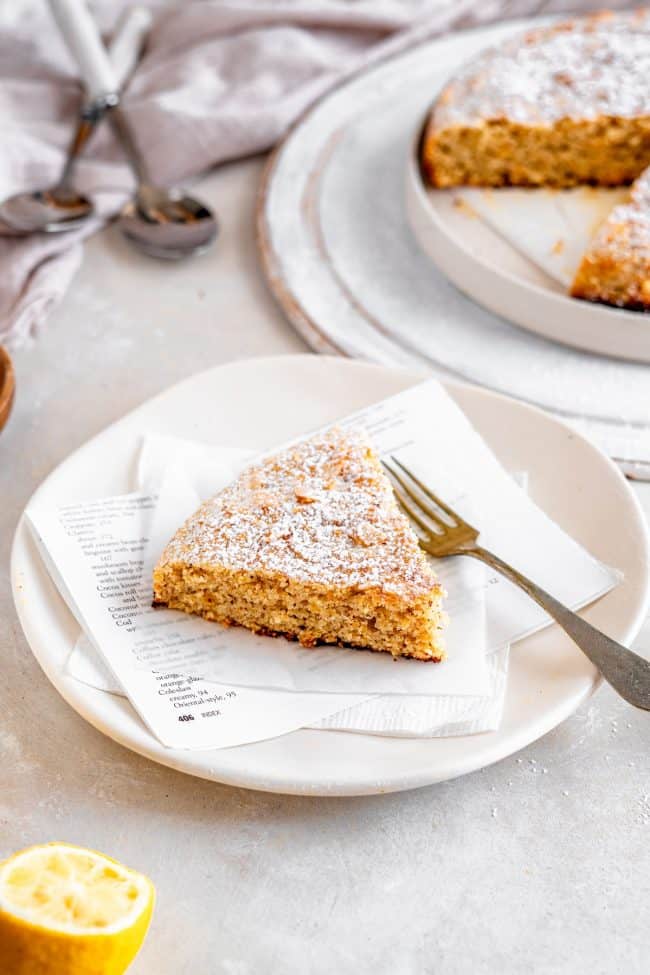 A single piece of Almond Cake on a white plate with a fork. Lemon and the rest of the cake surround it.